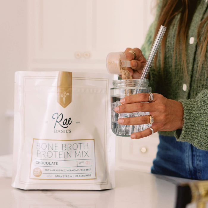 A scoop of Rae Basics Classic Chocolate Bone Broth Protein Mix going into a glass jar beside the bag
