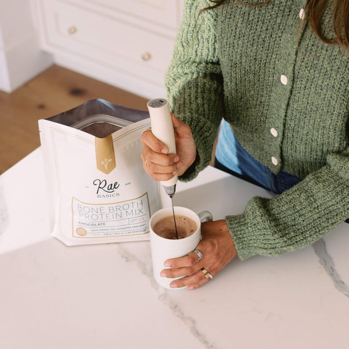 Woman frothing a mug of protein mocha with a handheld frother next to a bag of Rae Basics Bone Broth Protein Mix