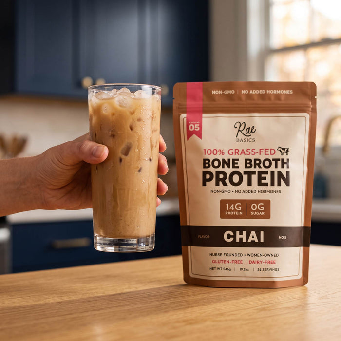 Hand holding a glass of iced chai next to a bag of Rae Basics Chai Bone Broth Protein Mix