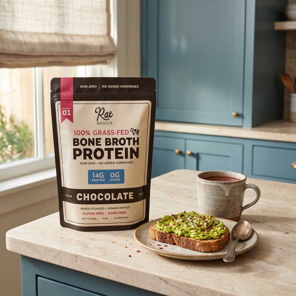 Bag of Rae Basics Classic Chocolate Bone Broth Protein Mix beside a mug and avocado toast on a kitchen counter