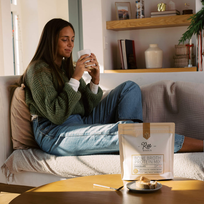 Woman relaxing on the couch with a warm mug beside a bag of Rae Basics Classic Chocolate Bone Broth Protein Mix