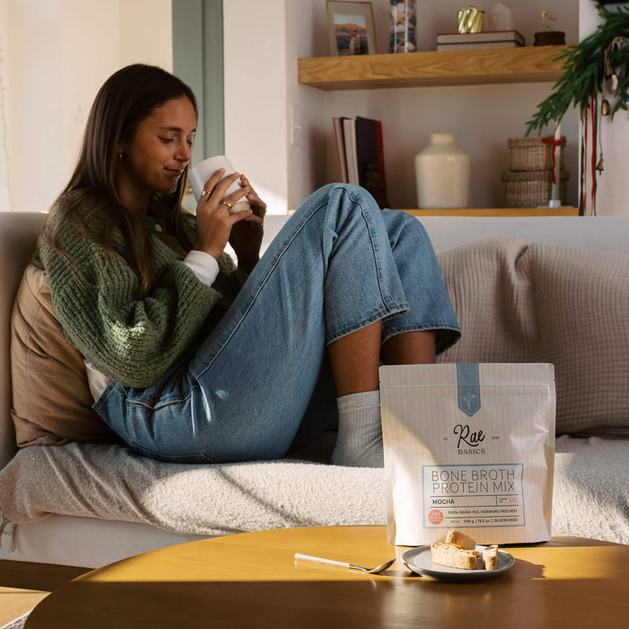 Woman relaxing on the couch with a warm mug beside a bag of Rae Basics Bone Broth Protein Mix