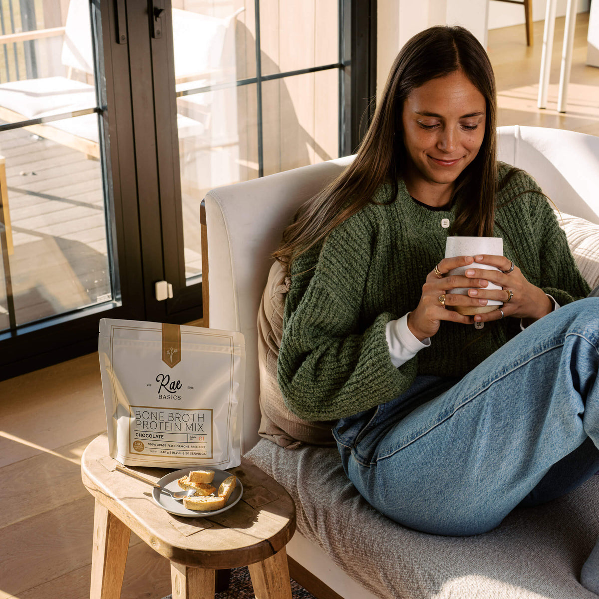Woman in a green cardigan smiling over a warm mug beside a bag of Rae Basics Bone Broth Protein Mix