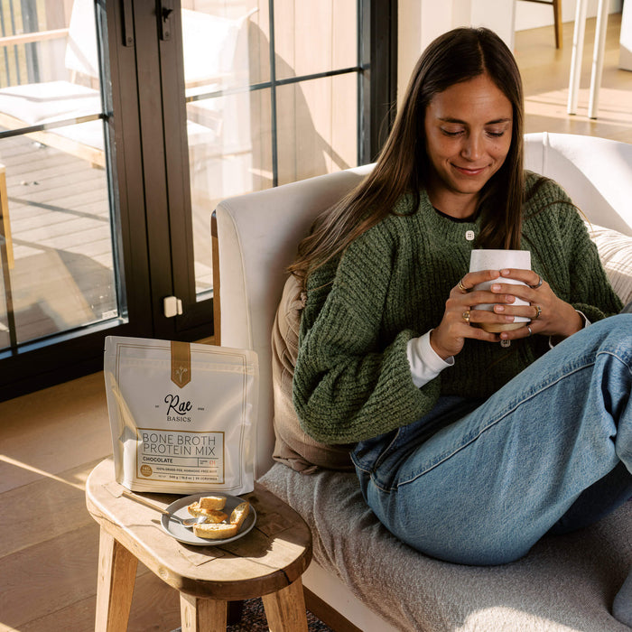 Woman in a green cardigan smiling over a warm mug beside a bag of Rae Basics Bone Broth Protein Mix