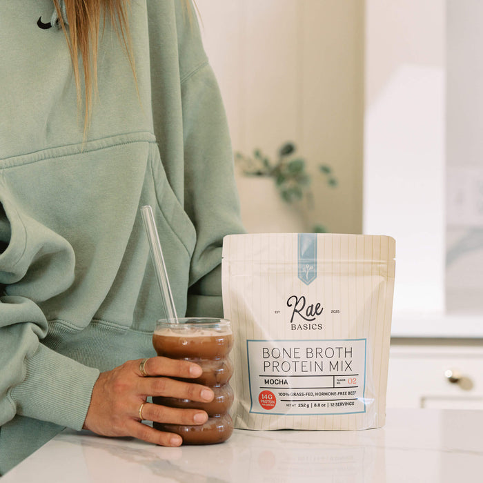 Woman holding an iced mocha next to a bag of Rae Basics Mocha Bone Broth Protein Mix on a marble counter