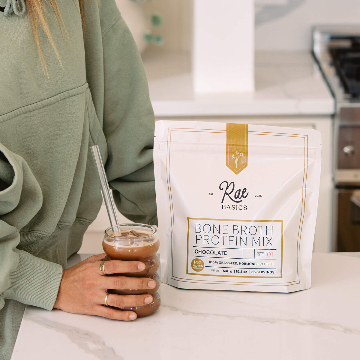 Woman holding an iced chocolate drink with a glass straw beside a bag of Rae Basics Classic Chocolate Bone Broth Protein Mix