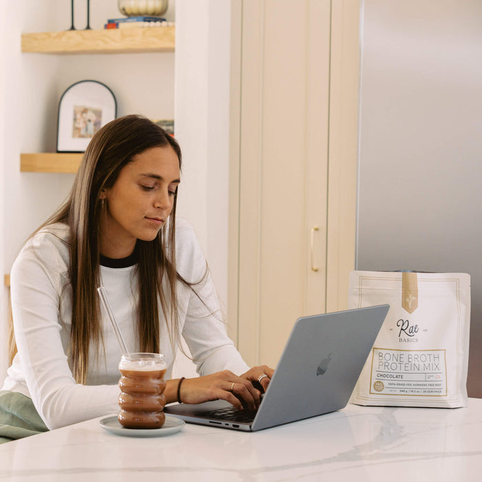 Woman working on a laptop at a kitchen counter with an iced mocha and a bag of Rae Basics Bone Broth Protein Mix