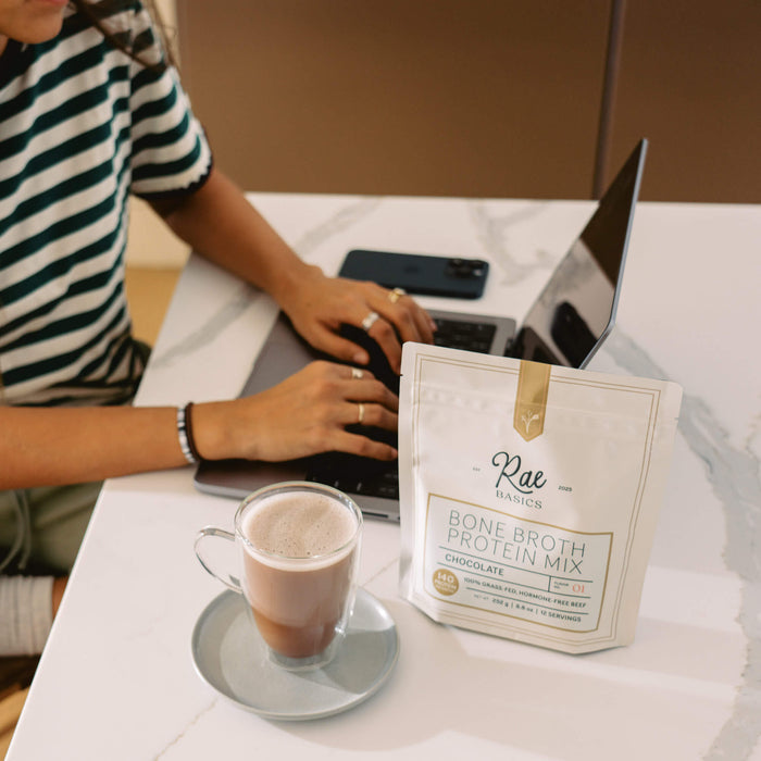 A glass mug of bone broth hot chocolate on a saucer beside a bag of Rae Basics Classic Chocolate Bone Broth Protein Mix