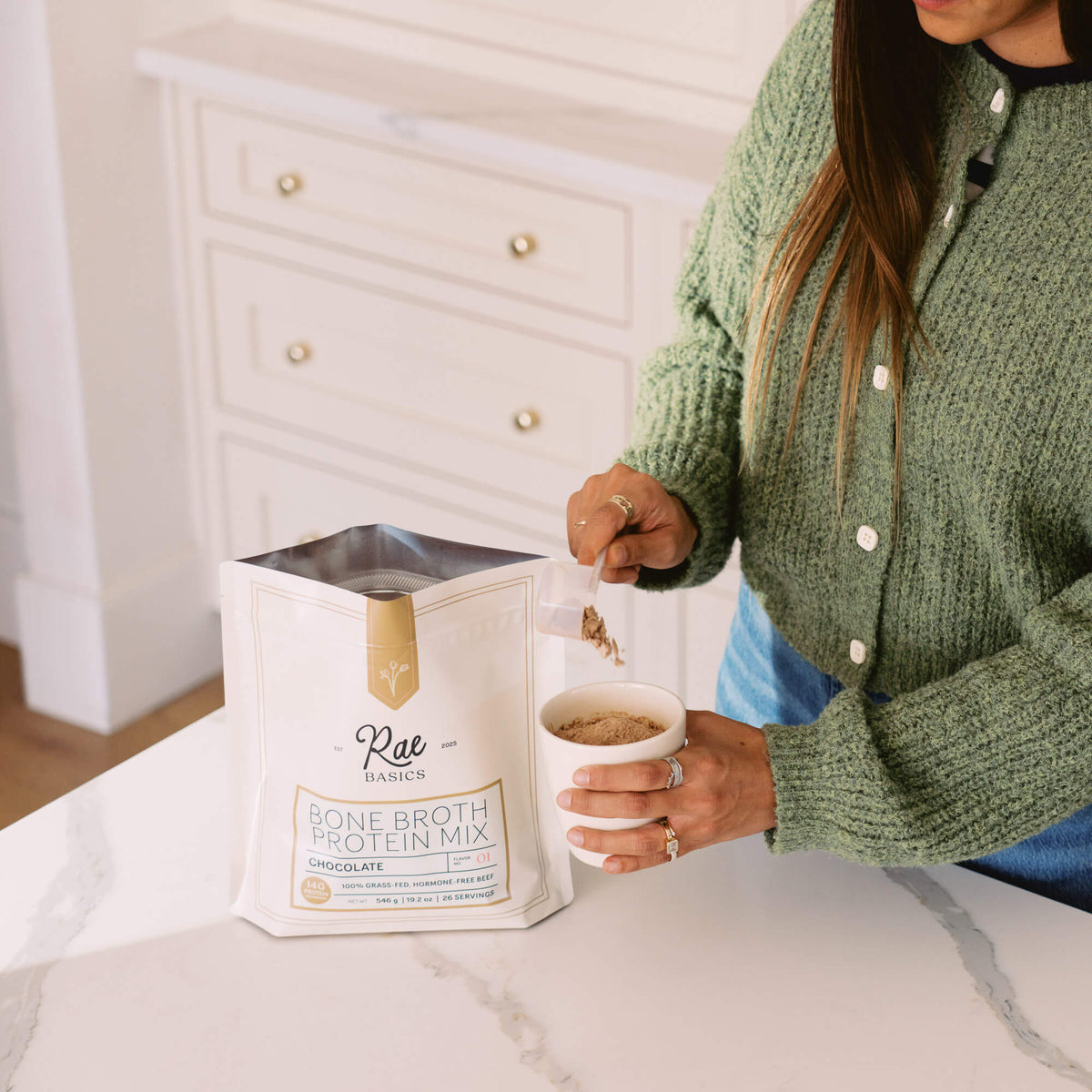 Woman spooning Classic Chocolate Bone Broth Protein Mix from the bag into a mug on a marble counter