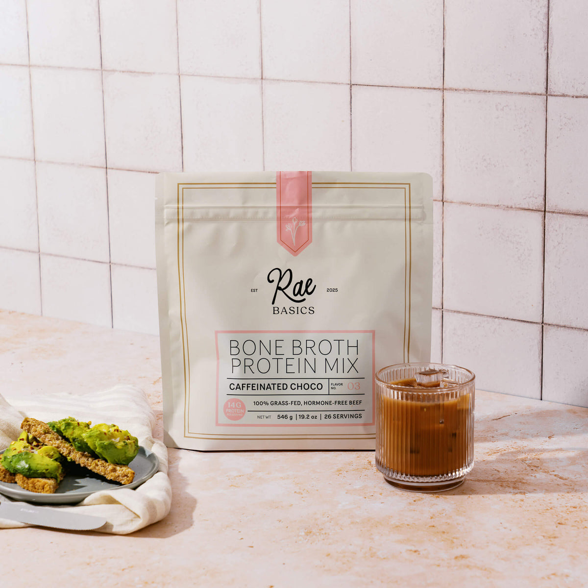 Bag of Rae Basics Caffeinated Chocolate Bone Broth Protein Mix beside an iced mocha and avocado toast on a kitchen counter