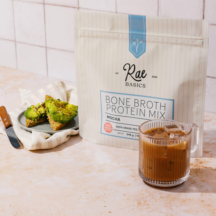 Iced mocha in a glass mug beside a bag of Rae Basics Mocha Bone Broth Protein Mix and avocado toast