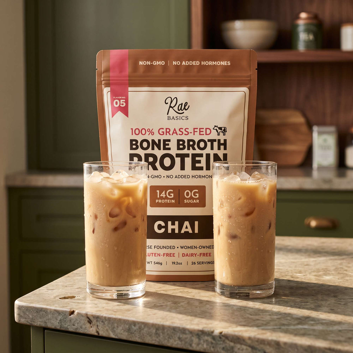 Two glasses of iced chai latte in front of a bag of Rae Basics Chai Bone Broth Protein Mix on a kitchen counter