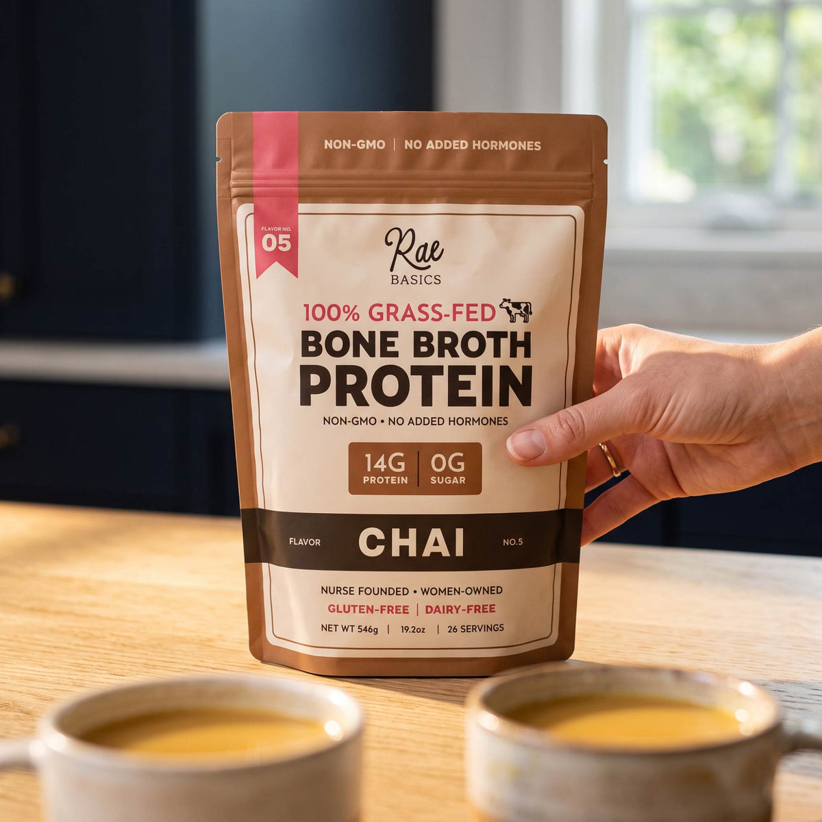 Hand holding a bag of Chai Bone Broth Protein Mix above two cups of warm chai latte