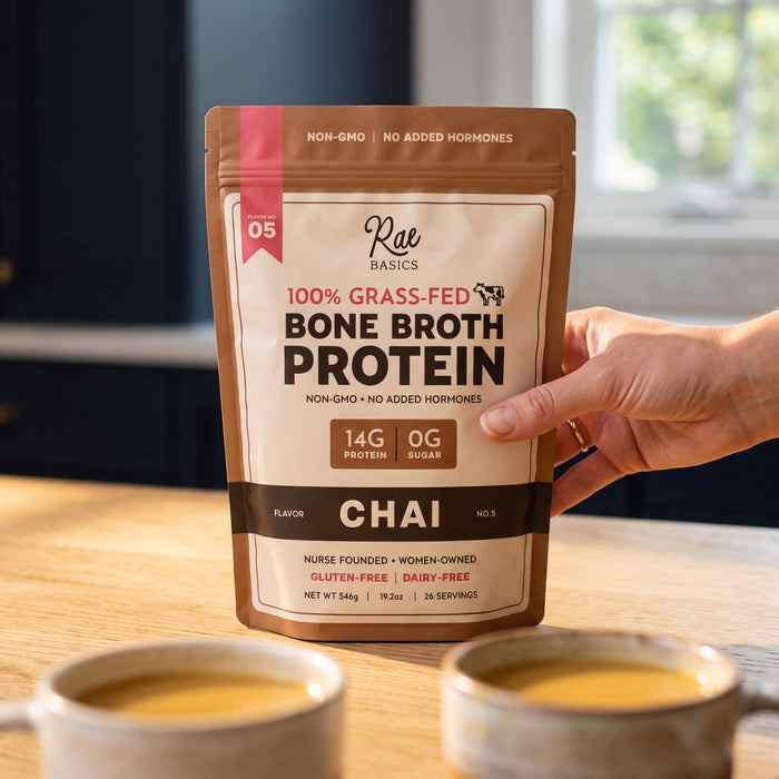 Hand holding a bag of Chai Bone Broth Protein Mix above two cups of warm chai latte
